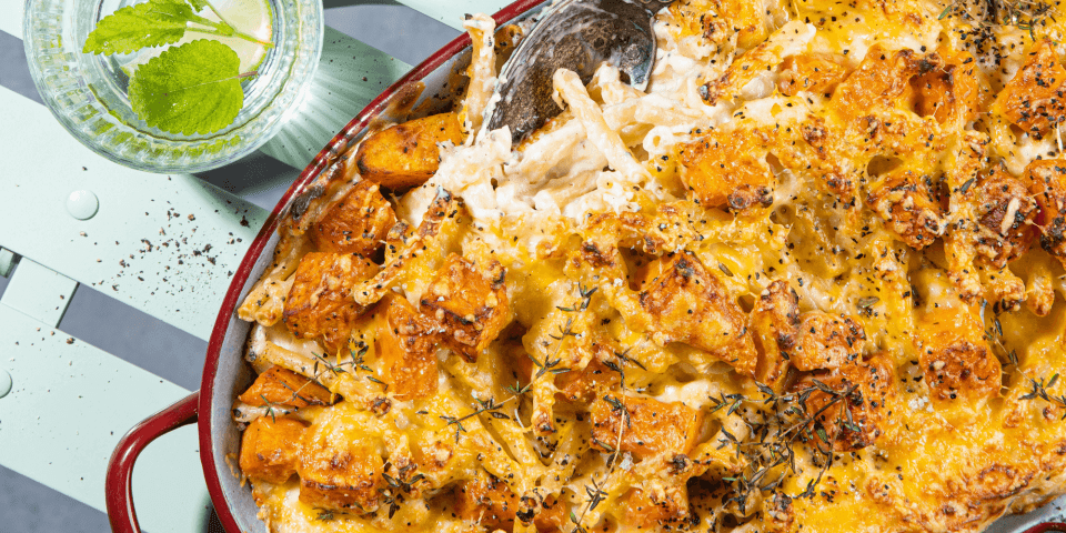 Mac en cheese met zoete aardappel Een grote bordeaukleurige ovenschotel staat op een licht blauwe stoel. De ovenschotel met ma&cheese en zoete aardappel is oranje kleurig. Boven de ovenschotel staan twee glaasjes water met ijsblokjes en verse munt.