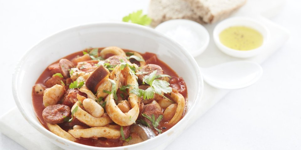 Mediterraans stoofpotje van inktvis en chorizo Een goed gevuld wit bord met het stoofpotje staat klaar om aan te vallen. De kleur is rood van de vele tomaten en chorizo die zijn gebruikt. Het bordje staat op een marmere snijplank waar ook brood en een klein potje olijfolie op staat.