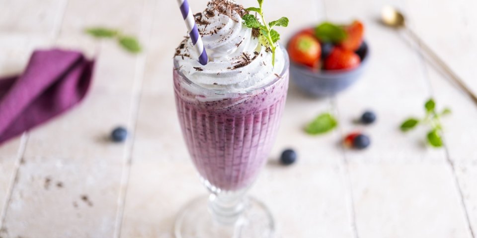 Milkshake van blauwe bes en aardbeien Een heerlijke grote milkshake staat op het aanrecht te wachten. Hij is mooi paars van kleur door de blauwe besjes gemixt met aardbeien. Er liggen enkele bessen op het aanrecht. De milkshake werd afgewerkt met een toef slagroom, muntblaadje en chocoladeshilfers. Ten aanval!