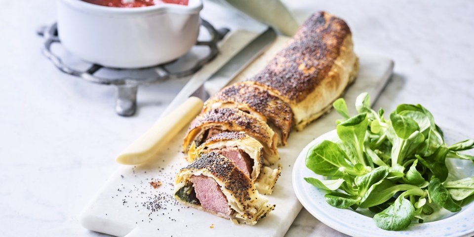 Mini beef wellington met spinazie en tomatensaus Een rol bladerdeeg ligt op een marmere snijplank. Het bladerdeeg is al aangesneden en zo zie je de spinazie en het vlees zitten. Wij serveren hem in kleine stukjes, net zoals een sushi, met homemade tomatensaus.