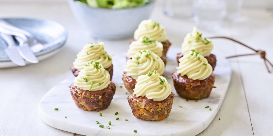 Mini-gehaktbroodjes met toefjes aardappelpuree Gehaktbroodjes die klaargemaakt zijn in een muffinvorm staan recht op een marmer snijplankje. Ze zijn afgewerkt met een toefje aardappelpuree dat er werd opgespoten. Ze zijn een feest om naar te kijken.