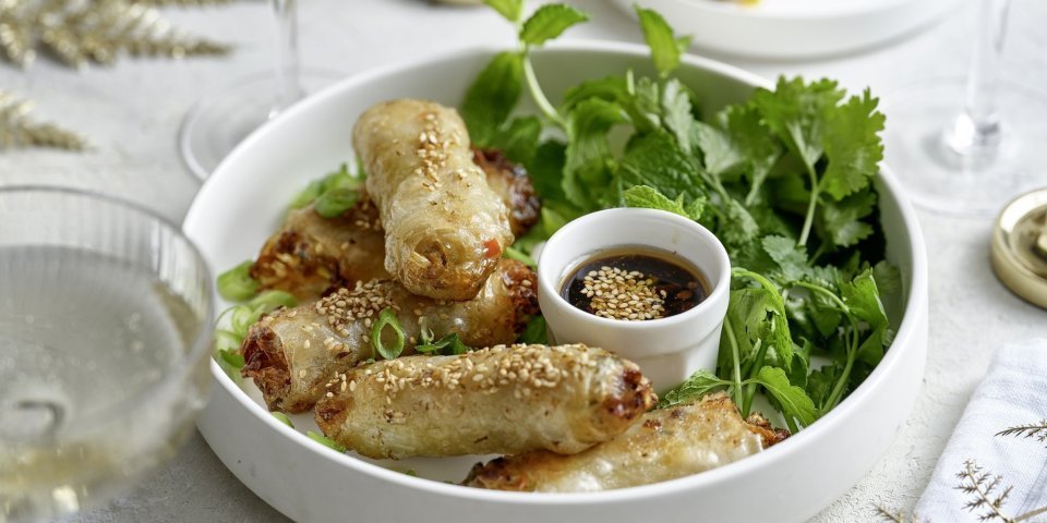 Mini-loempia met varkensvlees In een wit bordje liggen klaargemaakte miniloempia's, ideaal fingerfood. Ze liggen op een bedje verse kruiden en werden geserveerd met een dipsausje op basis van sojasaus. De tafel is gezet in feeststemming met mooie glaasjes en stoffen servetten.