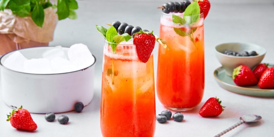 Mocktail van aardbei en basilicum Twee mocktails staan op het aanrecht, klaar om te drinken. Hij heeft een mooie rode kleur door de aardbeien, het glaasje is afgewerkt met ijsblokjes, takje munt, aardbei aan het glas en een spiesje met blauwe bessen opgeprikt.