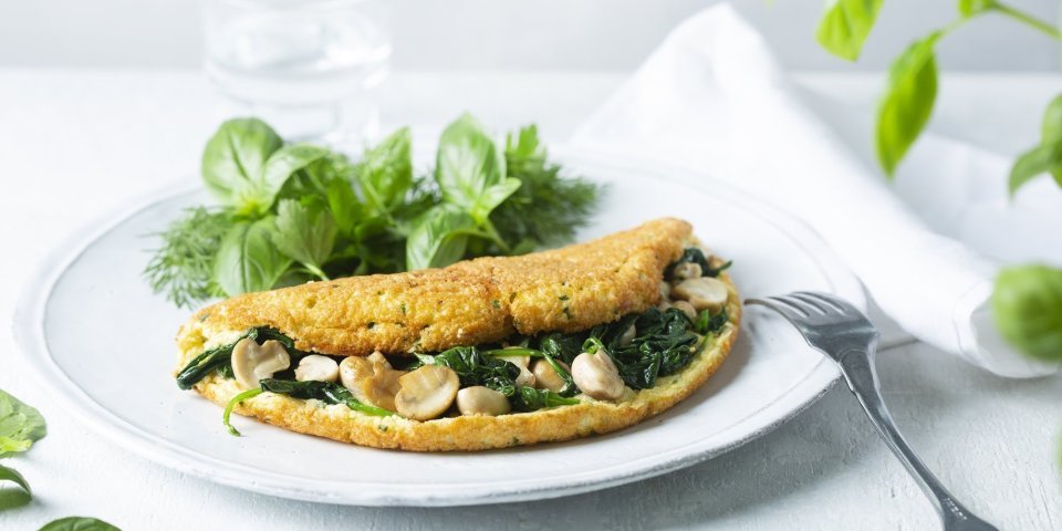 Een heerlijke dichtgevouwde omeletsoufflé gevuld met champignons, spinazie en kaas ligt op een wit bord. Het gerechtje werd afgewerkt met een slaatje van verse kruiden, denk maar aan basilicum en dille.  