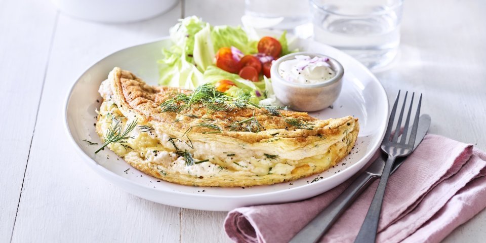 Omelet soufflé met verse kruiden en kaas Een heerlijke omelet ligt op een wit bord. Het is een luchtige omelette die we combineren met verse kruiden en kaas. We voegde ook nog wat sla en tomaatjes toe aan het bord voor een extra portie kleur en groenten.