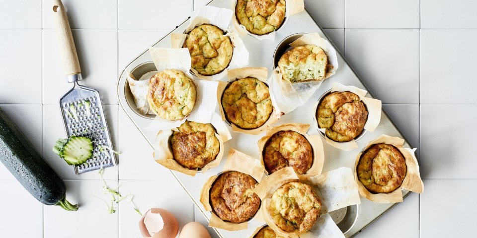 Ontbijtmuffins met courgette, yoghurt en Belgische kaas in een grote muffinvorm. Klaar om eruti te halen en op te eten. Naast de bakvorm ligt een verse courgette en lege eierschalen voor de sfeer.