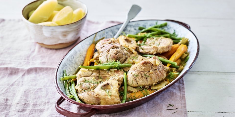 De kalfsschenkels liggen in een ovenschotel met groene boontjes en oranje wortels erbij. Dit zorgt voor een mooie variant op de klassieke ossobuco.