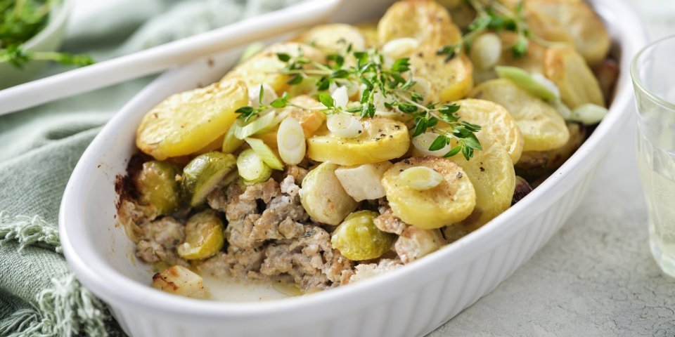 Ovenschotel met gehakt, groenten en aardappelschijfjes Een ovenschotel staat op het midden van de tafel, er is al een portie uitgeschept. Je ziet het gehakt onderaan liggen, met spruitjes , stukjes knolselder en goudgele aardappelschijfjes.
