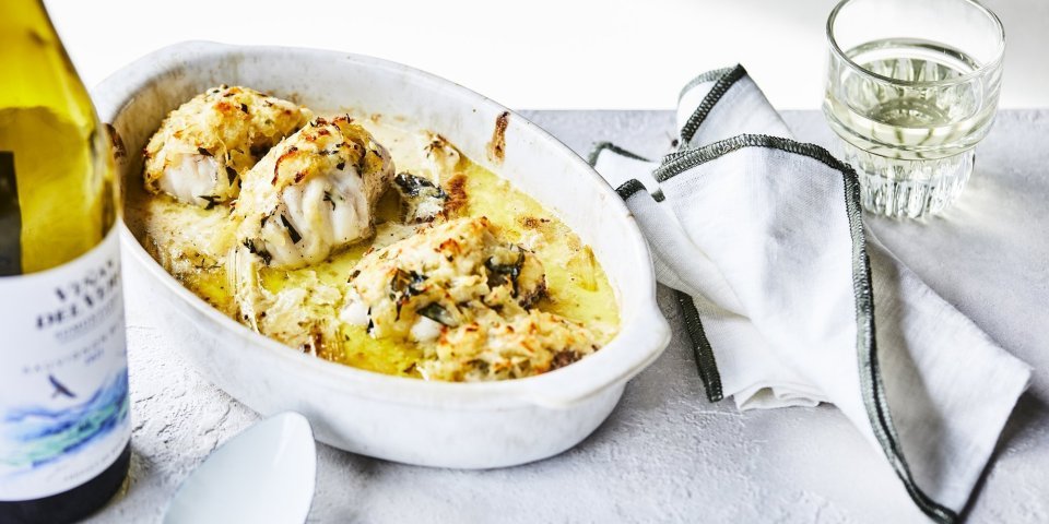 Een ovenschotel staat klaar op tafel om meteen te beginnen. De schotel is gevuld met stukken zeeduivel en prei in een mosterdsausje. Er ligt een krokant korstje op van geraspte aardappel en kaas. Er staat een flesje wijn langs en een witte scheplepel.