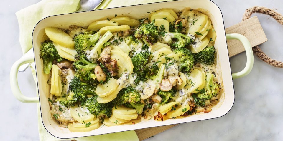 Een ovenschotel van aardappel, broccoli, champignons en blauwe kaas staat op het midden van het marmere aanrecht op een houten snijplank. Verse kruiden zorgen voor een extra groene touch!