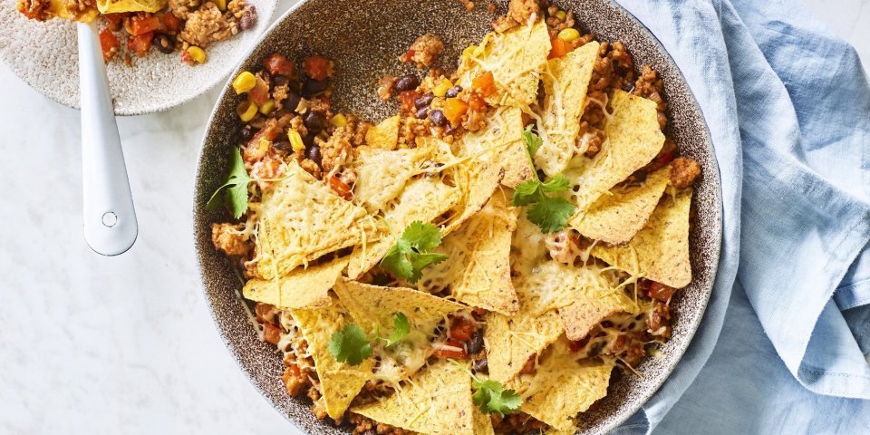 Ovenschotel van chili con carne met nachos en kaas Onderaan de ronde ovenschaal ligt de chili con carne met bovenop de nachos. Er is al een portie uitgeschept op een wit bordje en staat langs de ovenschaal. Er ligt ook een blauwe servette en enkele blaadjes kroiander voor de kleur.