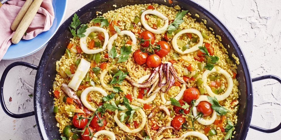 Paella van pijlinktvis met lente-ui, selder en kerstomaatjes Een grote pan gevuld met een kleurrijke paella staat op het aanrecht. Hij zit vol met de goudgele rijst, in ringen gesneden pijlinktvis, kerstomaten, paprika en verse peterselie.