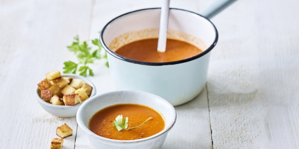 Op een houten witte tafel staat een kopje paprikasoep. De soepkom staat ook in beeld, dit is een lichtblauwe pot die mooi contrast geeft met de oranje-rode soep. De soep is afgewerkt met verse peterselie en croutons.