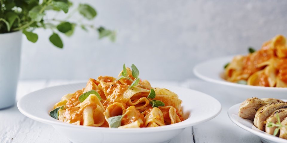 Pasta met geroosterde paprikasaus en kipfilet Een portie pasta met homemade paprikasaus staat klaar op een witte houten tafel. Een vers plantje oregano staat achteraan in beeld.