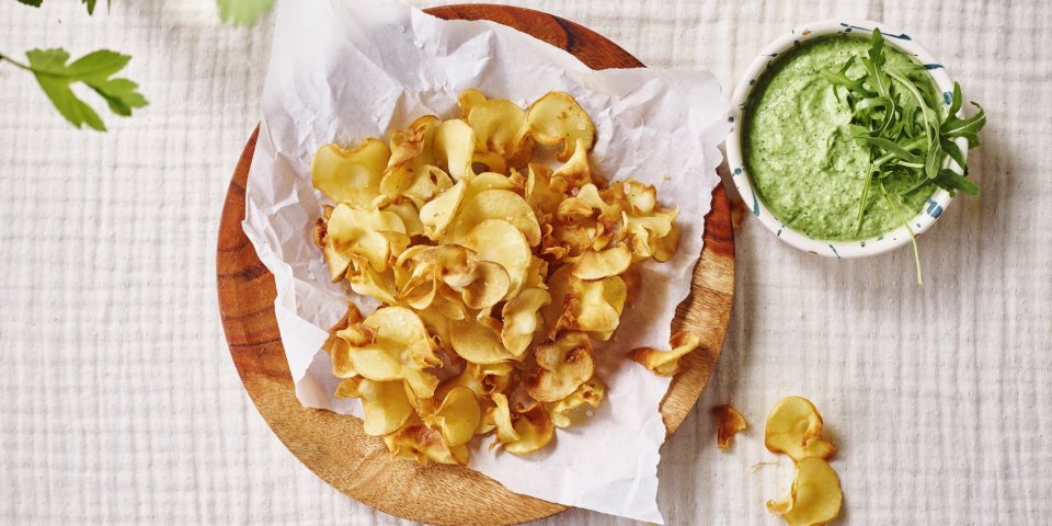 Pastinaakchips met een dip van broccoli en roomkaas Op tafel ligt een kalkwit tafelkleed, met hierop een houten bordje met wit boterpapiertje en chips van pastinaak op. Er staat een dippotje langs met de dip van broccoli en roomkaas.