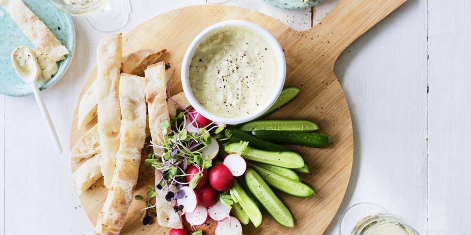 Pikante yoghurtdip met witte bonen en kruiden Op tafel staat een ronde houten snijplank met hierop een wit potje gevuld met onze pikante dip van yoghurt en witte bonen. Je kan de yoghurtdip gebruiken met stukjes snackkomkommer, brood en radijsjes in ons geval. Rond de plank staan enkele glazen witte wijn en helderblauwe broodbordjes.