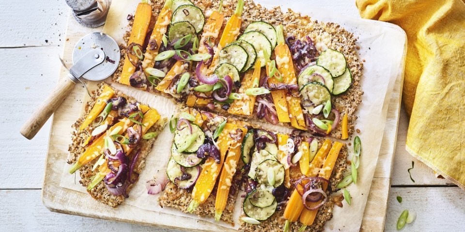 Plaattaart van bloemkool met uien, courgette en wortel Een krokante bodem van bloemkool en walnoten waarop kleurrijke groentjes liggen zoals wortel, courgetteplakjes en rode ui. De plaattaart ligt op een houten snijplank waar een pizzasnijder langs ligt, de pepermolen en goudgele servette.