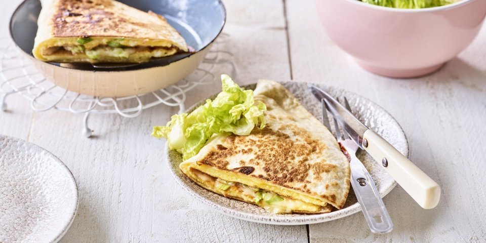 Een krokante tortilla ligt in een bordje klaar om aan te vallen. Hij is gevuld met een omelet, passendale kaas, gandaham en lente-ui. De tweede quesadilla ligt nog in de pan en staat ook op de tafel. 