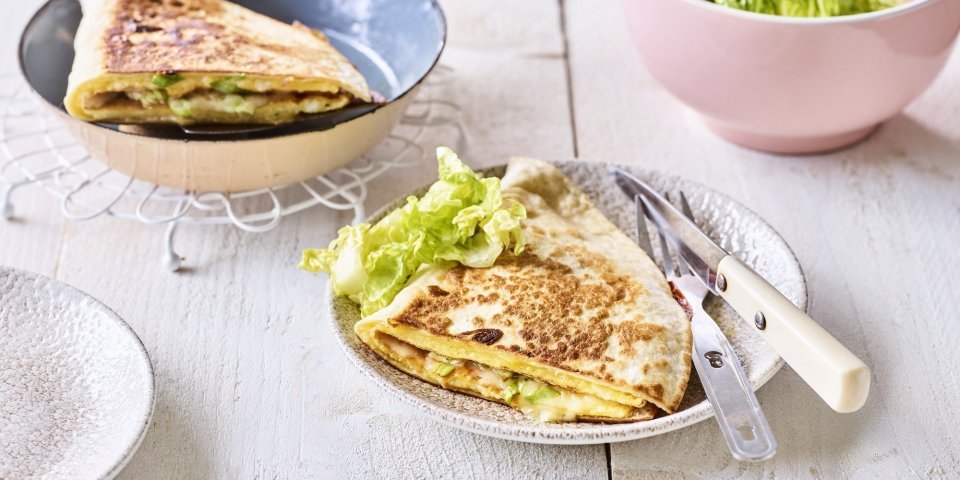 Quesadilla met een omelet en Passendale kaas Een krokante tortilla ligt in een bordje klaar om aan te vallen. Hij is gevuld met een omelet, passendale kaas, gandaham en lente-ui. De tweede quesadilla ligt nog in de pan en staat ook op de tafel.