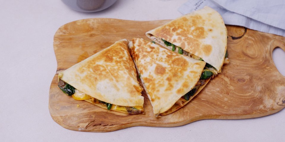 Een klaargemaakte quesadilla ligt op een houten snijplank. De quesadilla is in 3 gesneden, hij is gevuld met vlees, spinazie, zwarte bonen en kruiden. 