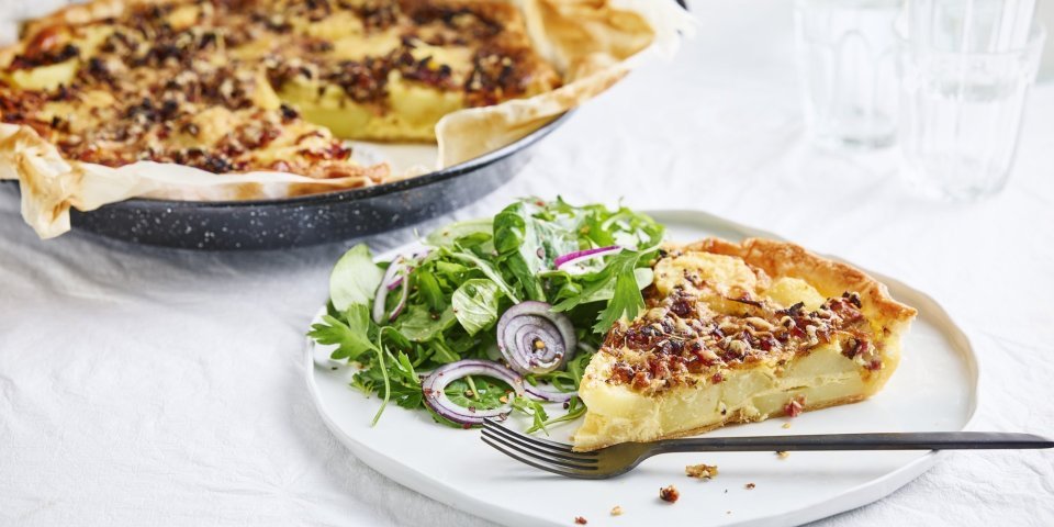 Quiche met aardappel, ui, spek en rozemarijn Er staat een quiche op tafel die aangesneden is. De quiche is gemaakt van schijfjes aardappel, ui, spek en rozemarijn. Wij serveren hem met een fris slaatje van kruiden op het bord, met een zoetzure dressing.