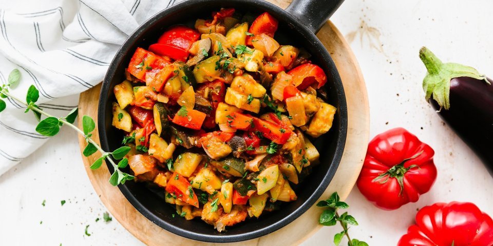 Ratatouille Op tafel staat de pan gevuld met homemade ratatouille boordevol groenten. Er liggen naast de gietijzeren pan twee rode tomaten en 1 aubergine.