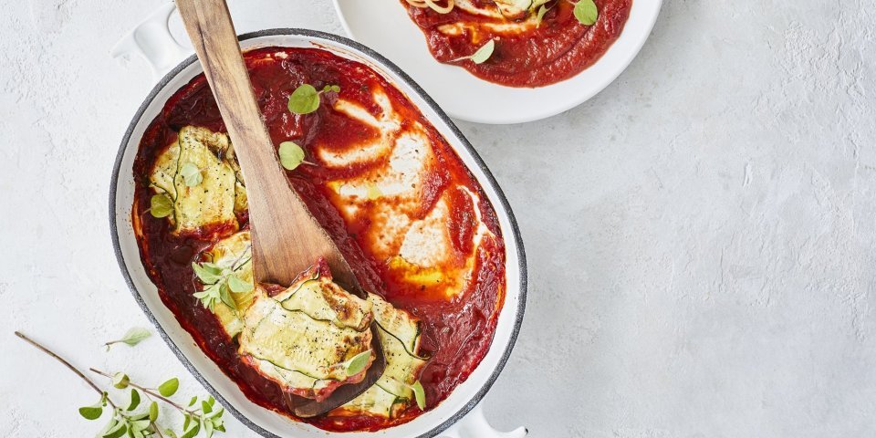 Pakjes van courgette liggen in witte ovenschaal met blauw randje. De tomatensaus ligt onder de ravioli van courgette, waardoor deze goed herkenbaar zijn. Er werd net een portie uitgeschept om meteen te eten. 