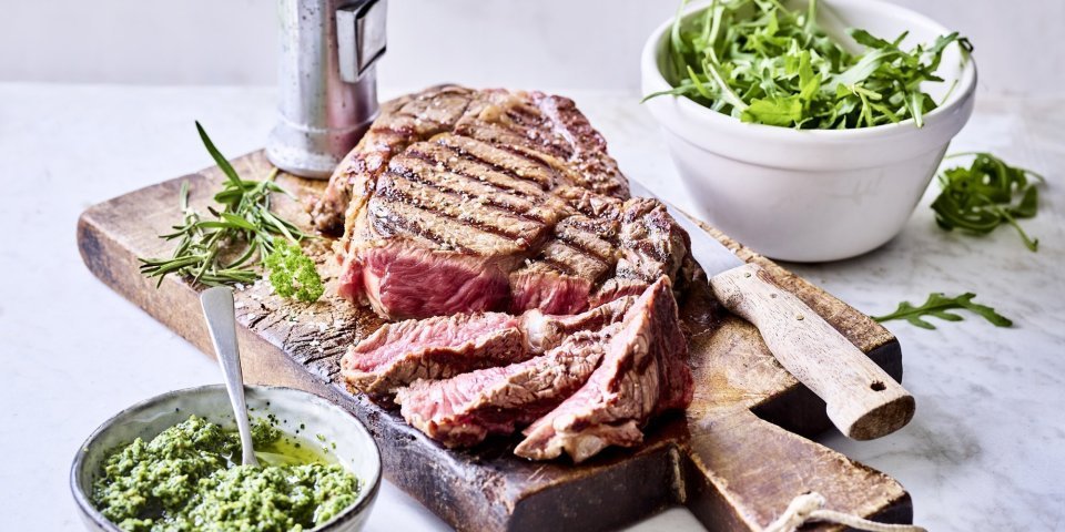 Ribeye steak op de barbecue met rucolapesto Een grote gegrilde steak ligt op een houten snijplank aangesneden. Er staat een potje homemade rucolapesto langs om meteen bij te scheppen bij je reepje vlees. Er staat ook een metalen pepermolen langs wie het graag wat pittiger wil.