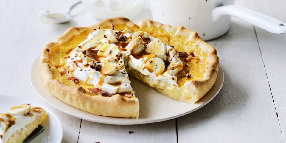 Rijsttaart met slagroom, banaan en gezouten karamel Op een witte houten tafel staat een klaargemaakte rijsttaart, hij is afgewerkt met vers opgeklopte slagroom, stukjes banaan en vloeibare karamel. Er is al een stukje aangesneden. Verder uit beeld staat het steelpannetje waarin de karamelsaus is klaargemaakt.