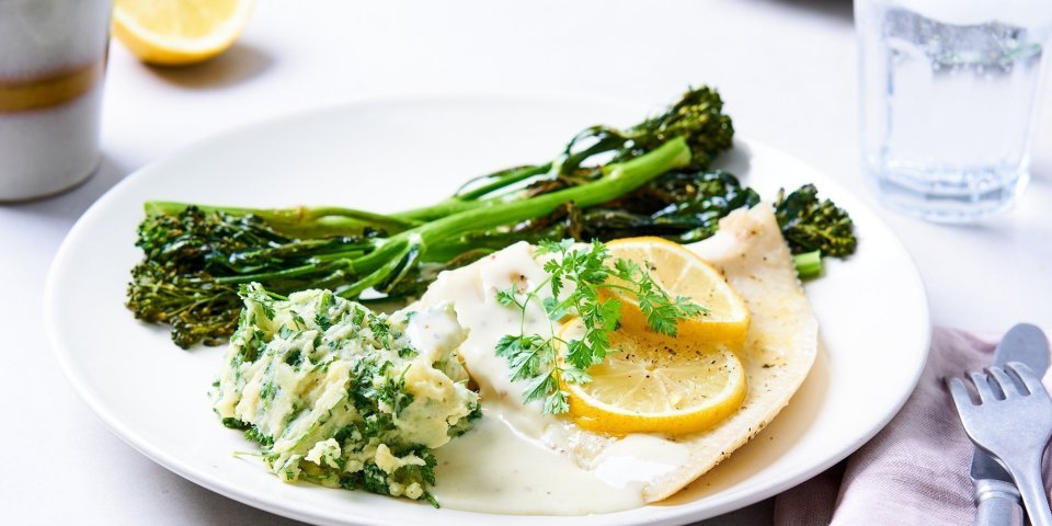 Rog uit de oven met mosterdsaus, bimi en kervelpuree Op tafel staat een wit groot bord met hierop de roggevleugel, citroenschijfjes, gegrilde bimi en een schep kervelpuree. Er staat een glas water langs, samen met een roze linnen servette en bestek.