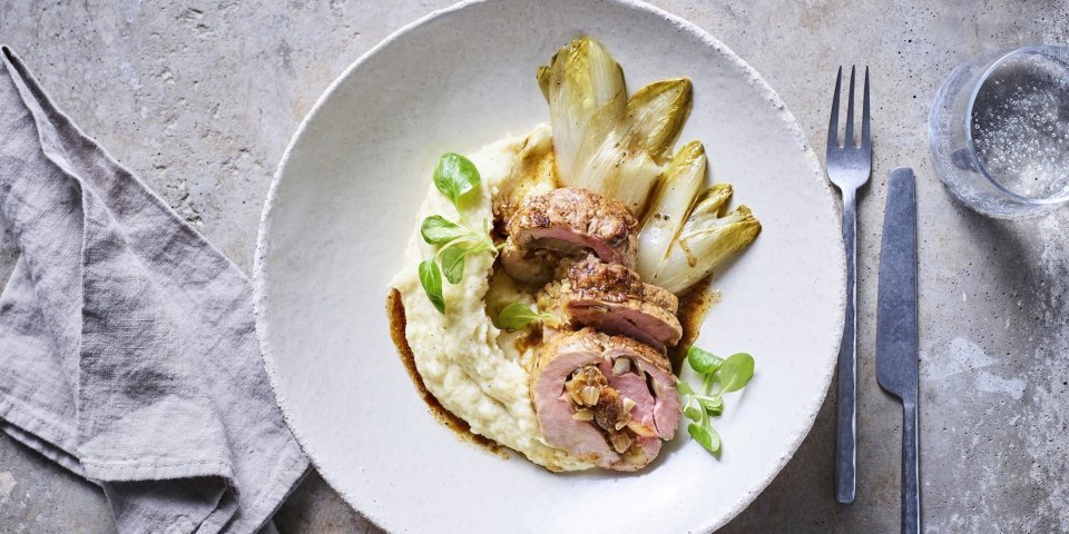 Rollade van varkenshaasje met witloof en pastinaakpuree Een stukje rollade van varkensvlees ligt in een wit groot bordje op een smeuige pastinaakpuree met gebakken witloofstronkjes.