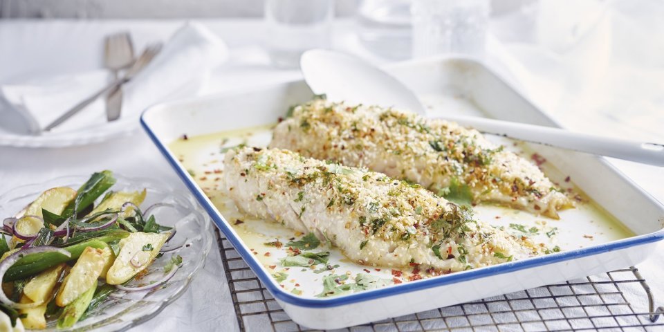 Roodbaars met een crumble van gember, sesam en chili met aardappelsalade De gegrilde roodbaars uit de oven ligt op een witte bakplaat en staat op een rooster. De crumble zorgt voor een zomerse sfeer in het gerecht. Naast de ovenschaal staat een bordje met wat aardappelsalade uitgeschept.