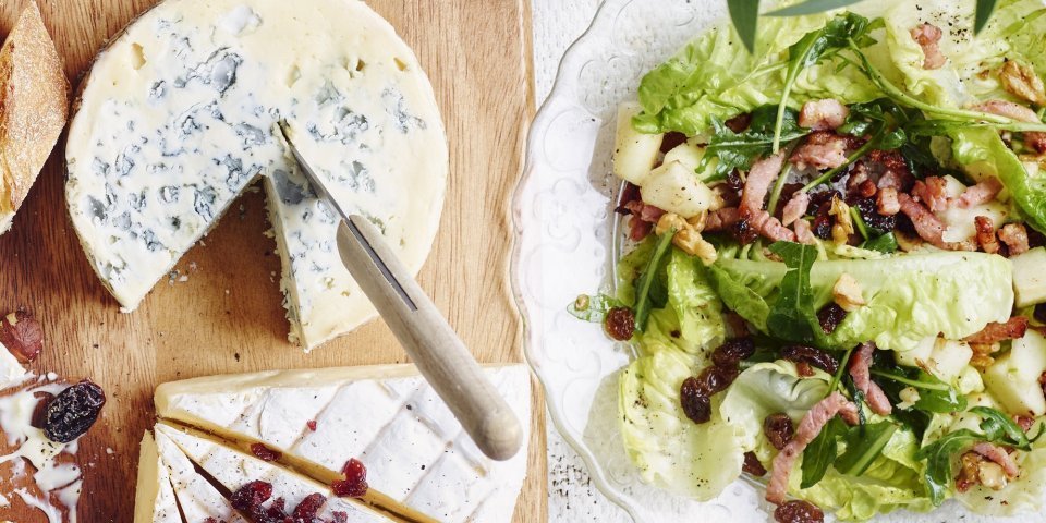 Een heerlijk salade met little gems, frisse appelstukjes en krokante spekblokjes staan op tafel in een glazen slakom. De slakom staat naast een kaasplank met een stuk blauwekaas en Belgische brie.