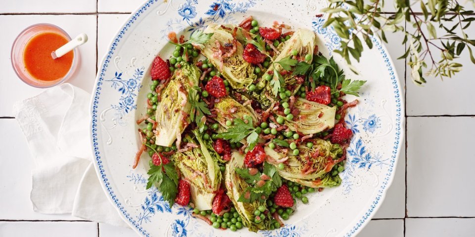 Salade met frambozen, spekjes en frambozendressing Een wit retro bordje staat op het aanrecht. Het is gevuld met een salade, gebakken spekjes en little gem. De frambozendressing is helder en roze, deze staat naast het bord in een glazen potje.