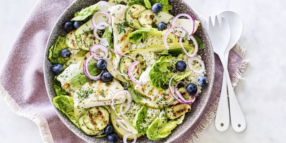 Salade met gegrilde zeetong, courgette en peer Een diep bord gevuld met sla, gegrilde zeetong, courgette, peer en blauwe bessen. Onder het bord ligt een paars servette en wit bestek om meteen dit gerechtje te verorberen.