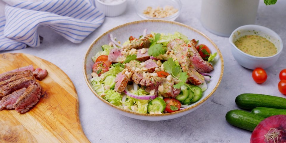 Een heerlijke saladekom is klaargemaakt en staat op het aanrecht nog te pronken. De bowl is rijkelijk gevuld met sla, Chinese kool, komkommerstukjes en tomaat. Hier bovenop liggen de reepjes steak die saignant gebakken zijn. De salade is afgewerkt met de dressing, de overige dressing staat in een potje naast de salade.. 