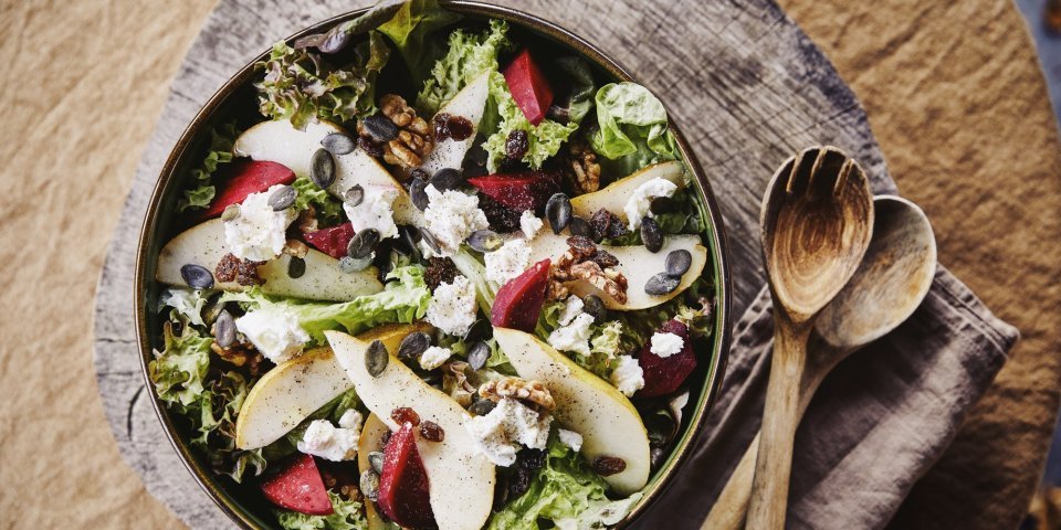 Een mooie slakom boordevol heerlijke producten. Deze side dish is gemaakt met eikenbladsla, rode bietjes, peer, walnoten en stukjes geitenkaas. Er ligt houten slabestek naast op een warmbruine linnen servette. 