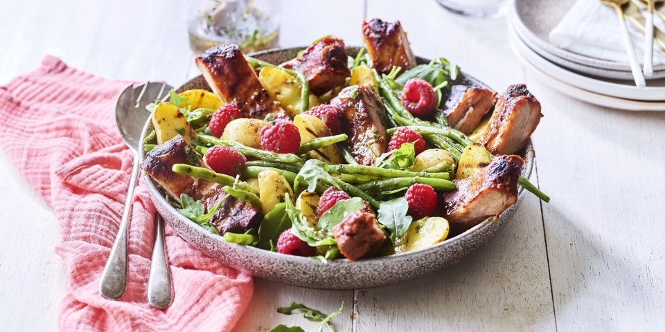 Salade van krieltjes, boontjes, frambozen en ribbetjes Een grote serveerschaal is gevuld met de aardappelsalade. Je ziet de framboosjes, boontjes en ribbetjes in de salade zitten. Er staat een torentje bordjes klaar met bestek om de salade te verdelen en op te eten.