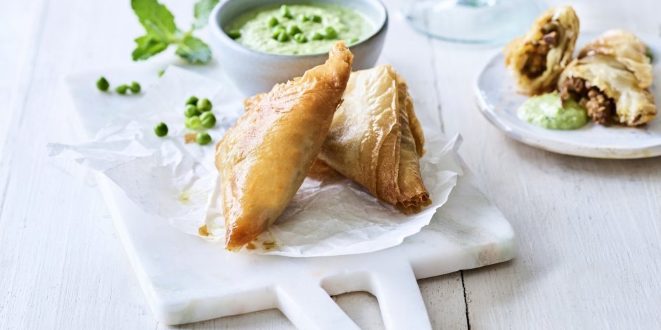 Klaargemaaakte samosa met lamsvlees liggen gepresenteerd op een marmer plankje. Ze worden opgediend met een frisgroene dip van munt en erwtjes.