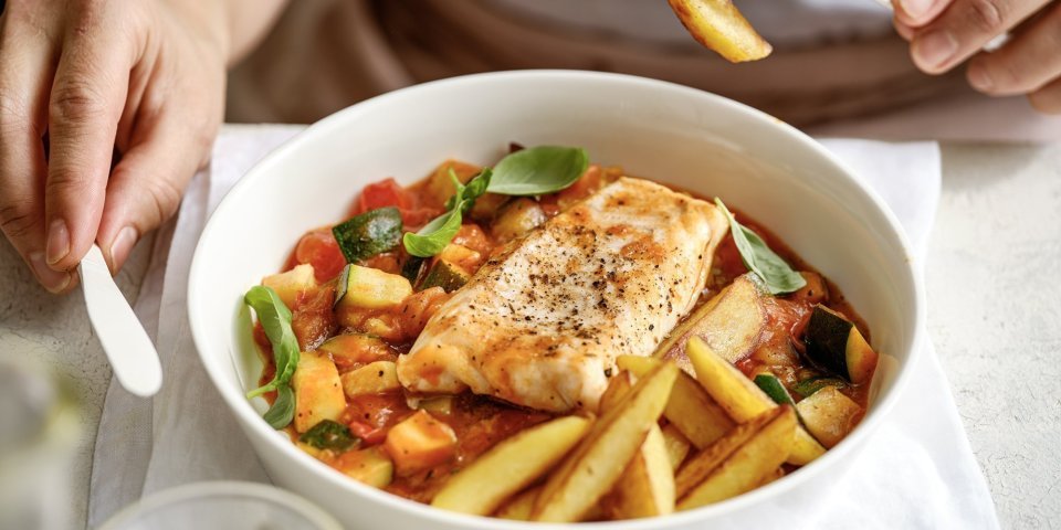 Aan taffel zit een vrouw die een stukje aardappel prikte op haar vork. Voor haar staat een bordje gevuld met het gerecht, vis met Prvençaalse saus en gebakken aardappelpartjes. 