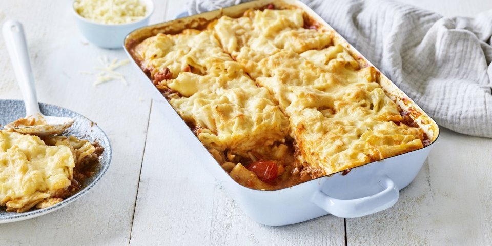 Een lichtblauwe ovenschotel staat op een houten witte tafel. De ovenschotel is gevuld met een mac & cheese én een tomatensaus, waardoor hij eerder weg heeft van een lasagne. Er is al 1 portie uitgeschept en wil al beginnen aan de volgende. Laat het niet koud worden. 