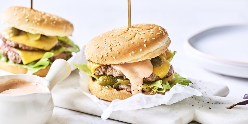 Smashburger met homemade bbq kruiden en kaas Twee heerlijke burgers liggen klaar op een marmere snijplank. Het vlees is verdeeld in twee dunne porties met de saus en gesmolten kaas er bovenop. De burgers worden recht gehouden door middel van een prikker.