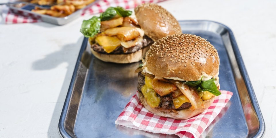 Twee hamburgers liggen op een inoxe plateau. De ene burger moet zijn hoedje nog krijgen.