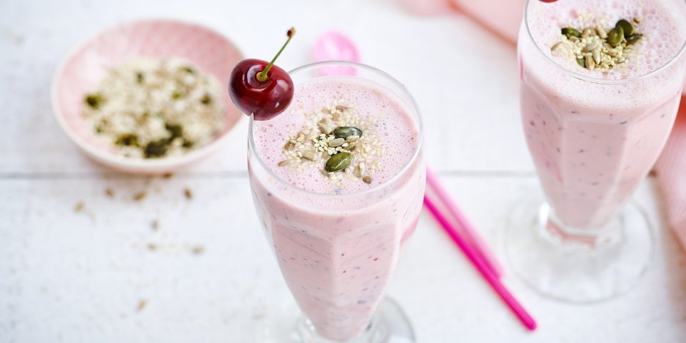 Twee rooskleurige smoothies op een witte houten tafel. De smoothie is afgewerkt met een kers en enkele pitjes en zaadjes. Een snel en makkelijk drankje met feestallures! 