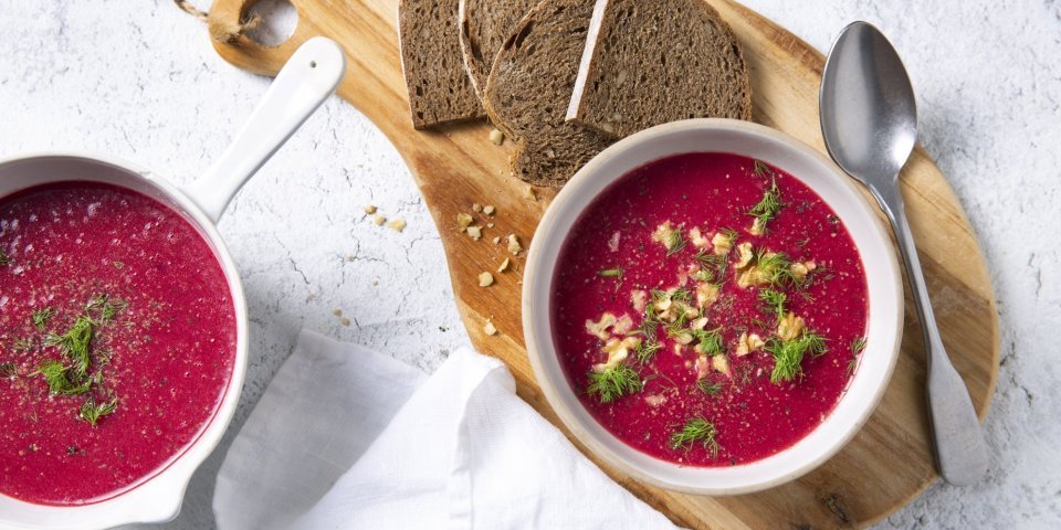 Soep van rode biet met aardappel, zure room en komijn Soep van rode biet kleurt natuurlijk rood en smaakt naar nog! Ze is geserveerd in twee witte kommetjes en staat op een houten snijplank met enkele bruine sneden brood bij.
