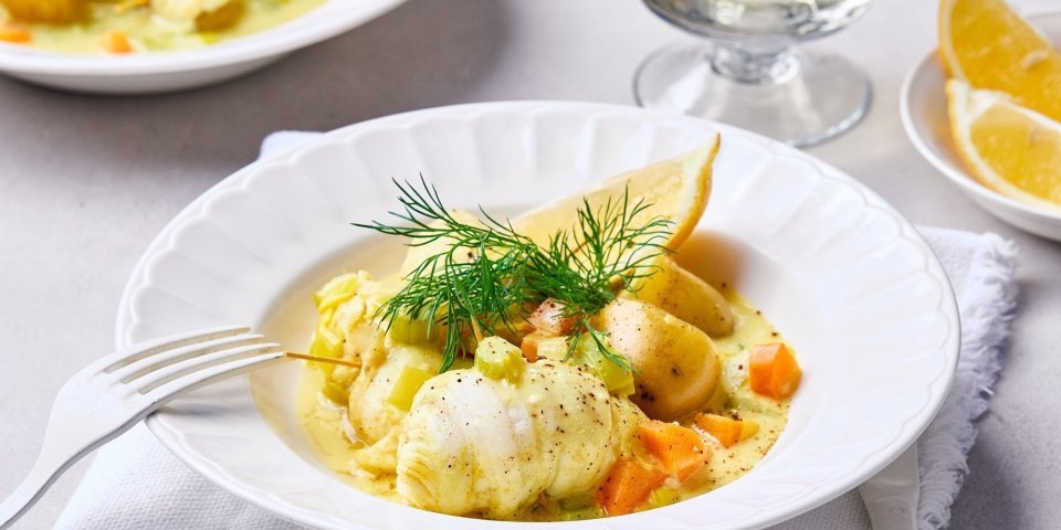 Een wit diep bord staat op de tafel, hij is gevuld met een geelkleurig stoofpotje van vis door de curry. Het visstoofpotje is afgewerkt met dille en een partje citroen.