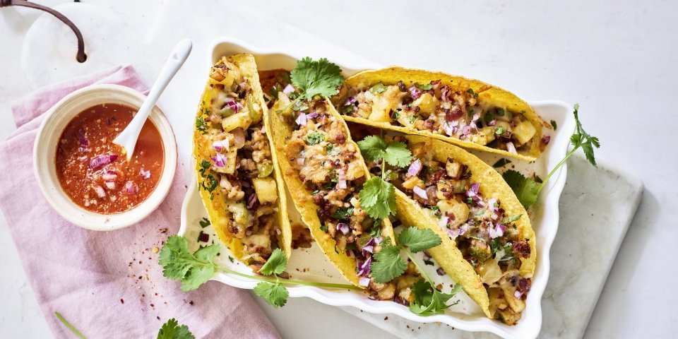 Taco’s met aardappelen en gehakt Vier tacos liggen klaar in een ovenschaal om aan te vallen. De pittige tomatendip staat links van de schotel op een roze servette. Het gerechtje is afgewerkt met verse korianderblaadjes.