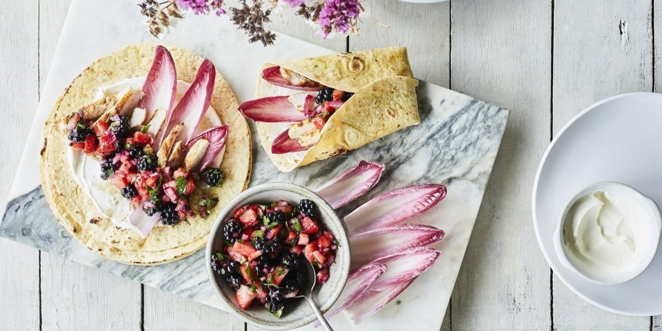 Taco’s met kip, roodlof en een salsa van braambessen en aardbei ligt klaar om op te eten. Er ligt al een tweede taco in de maak, op een marmere snijplank. De kleuren van roos tot donker rood van het fruit en loof geven een mooi contrast in de foto.