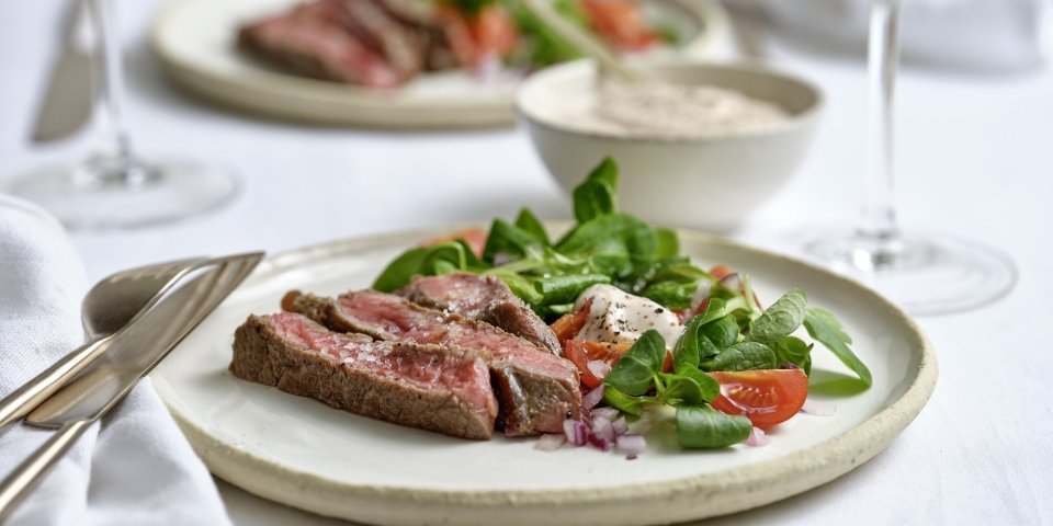 Tagliata van zesrib met veldsla en tomaat Twee borden zijn geserveerd op tafel, de licht beige borden zijn gevuld met reepjes gegrild vlees op een gemengde en frisse sla van veldsla en kerstomaten. Er staat ook een glaasje witte wijn op tafel.