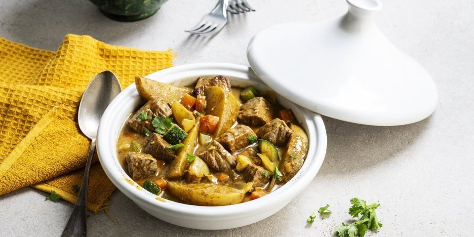Tajine met rundsvlees, aardappelen en groenten Op tafel staat een witte tajine met een gerechtje van rundvlees, courgette, selder, wortel en stukjes aardappel. Er ligt een okerkleurige servette bij en groene kommetjes om de tajine in op te scheppen.